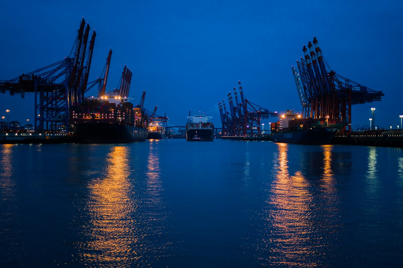 Ship-to-shore cranes and berthed container ships at blue hour, lights reflected in the harbour