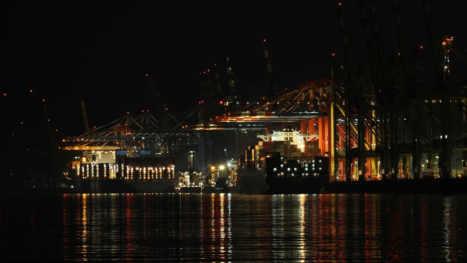 Container terminal cranes and a loaded ship lit up at night, reflected in the water