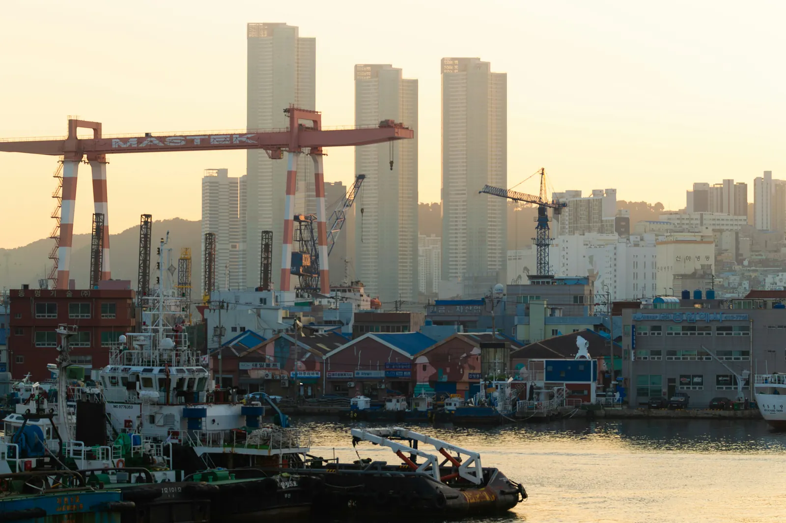 Busan harbour at golden hour: shipyard cranes, tugs and the city's towers