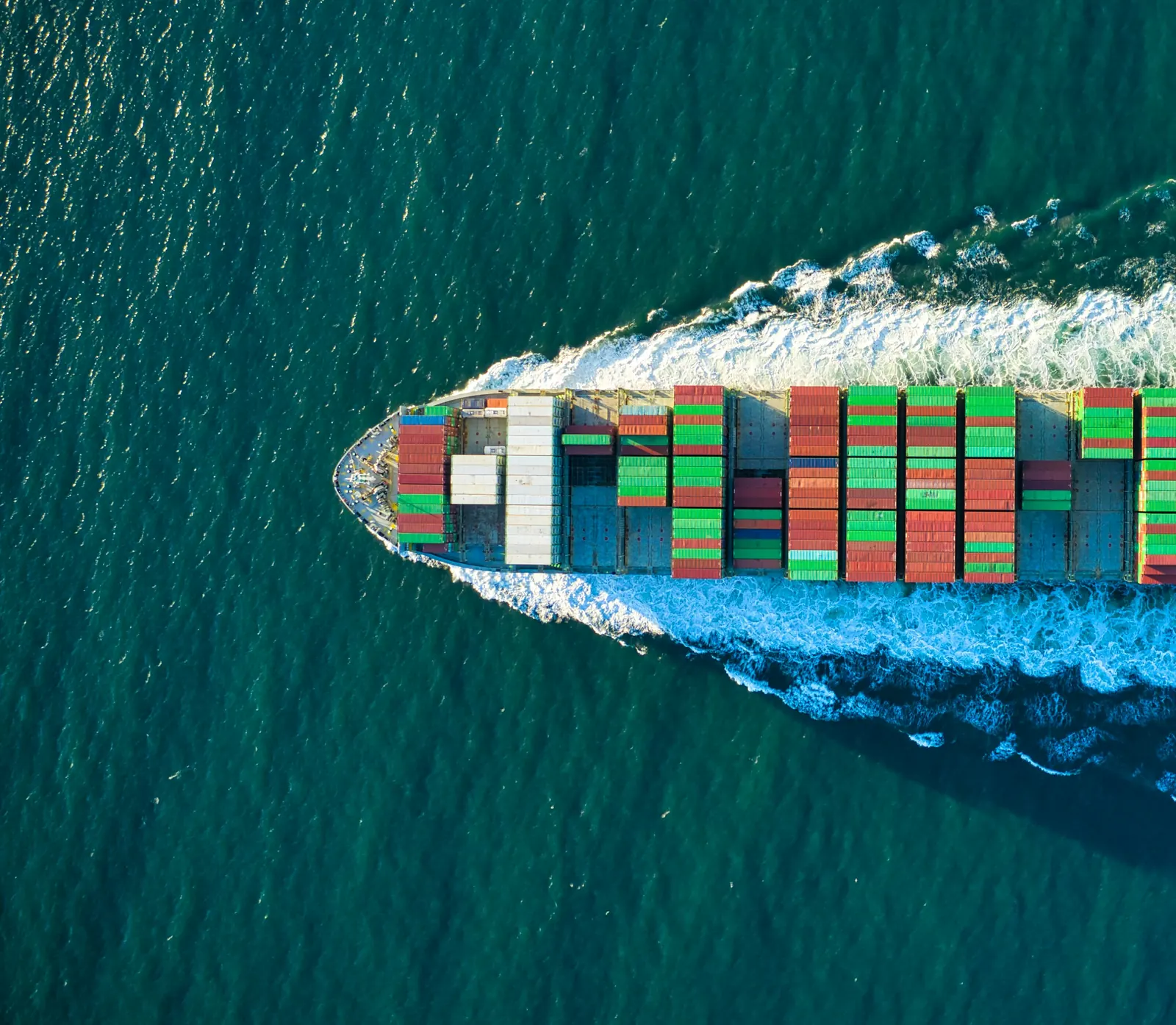 Aerial view of a loaded container ship cutting a white wake through green-blue sea
