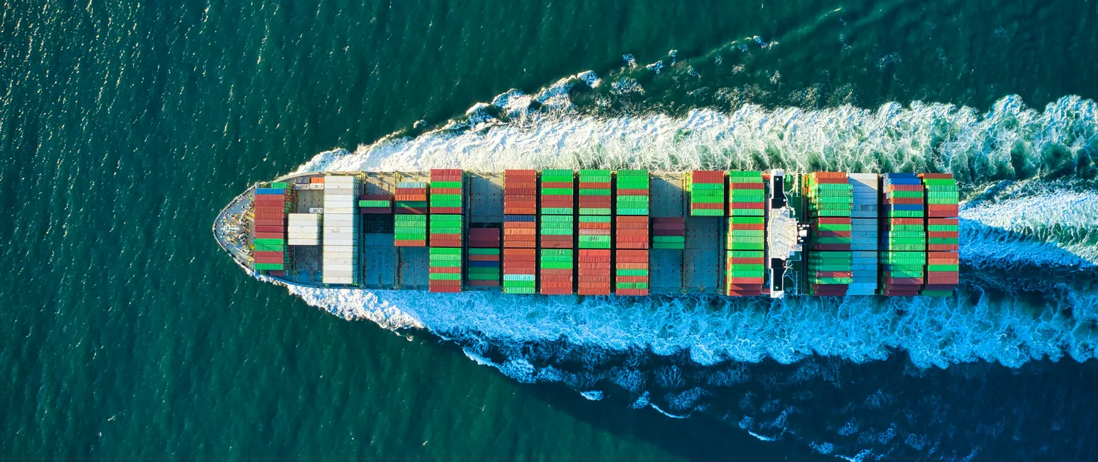 Aerial view of a loaded container ship cutting a white wake through green-blue sea